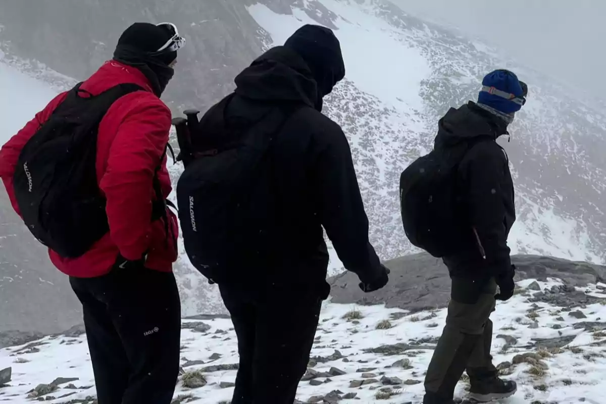 Tres personas con ropa de abrigo y mochilas caminan por un paisaje nevado y montañoso.