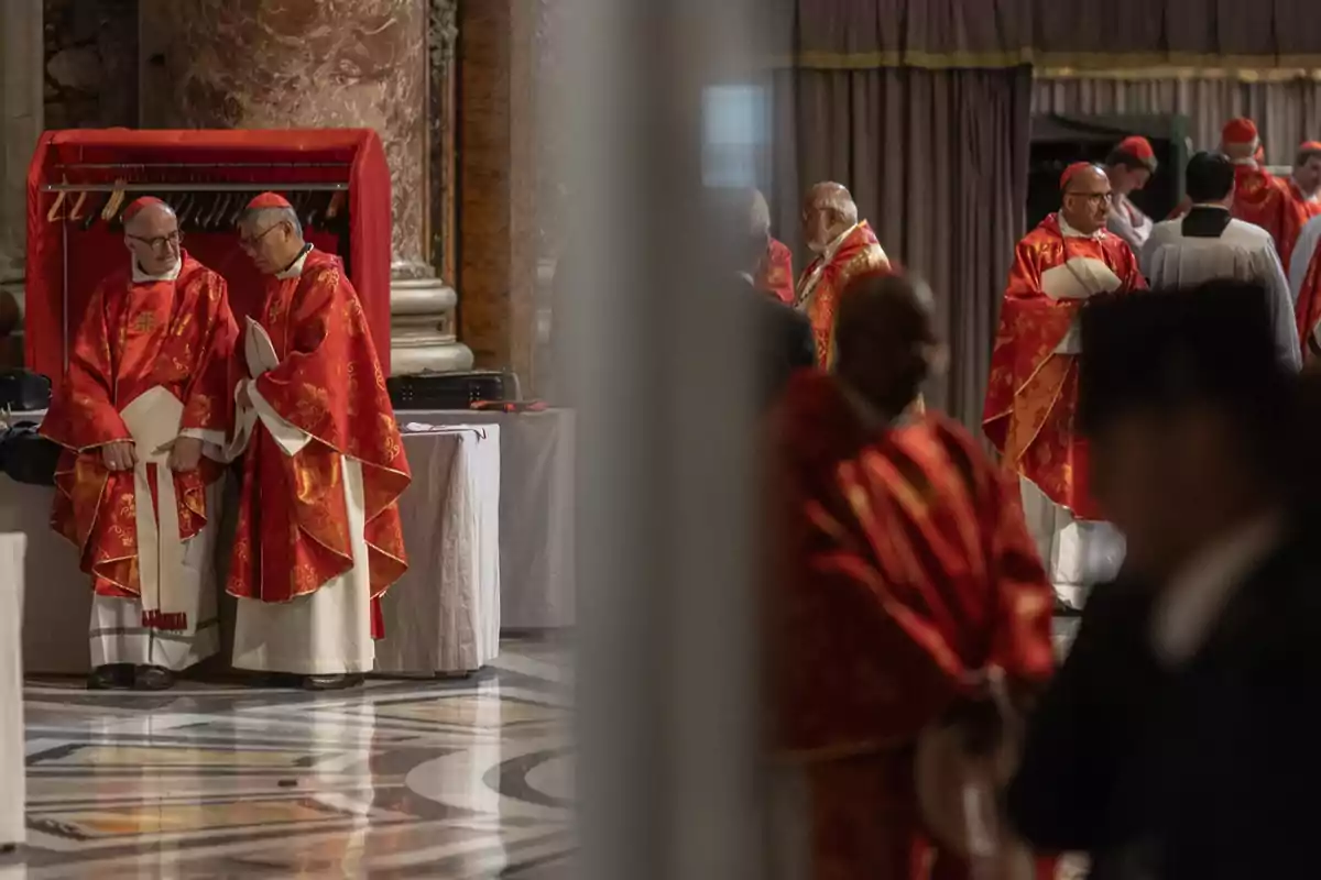 Un grupo de cardenales vestidos con vestimentas rojas se encuentra reunido en un espacio interior con columnas de mármol y cortinas, algunos conversan entre sí mientras otros están de pie en el fondo. Un grupo de cardenales vestidos con vestimentas rojas se encuentra reunido en un espacio interior con columnas de mármol y cortinas, algunos conversan entre sí mientras otros están de pie en el fondo.