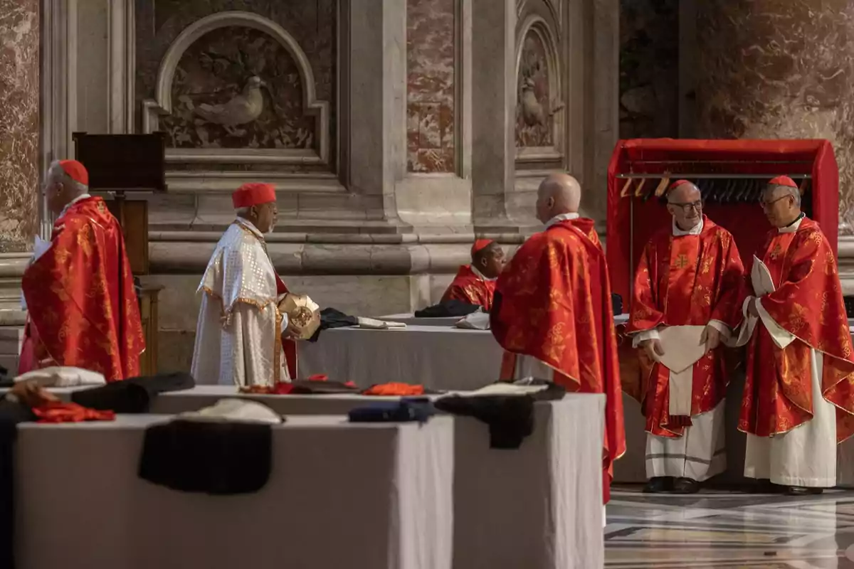 Un grupo de cardenales vestidos con vestimentas rojas se encuentra reunido en un espacio con decoración religiosa, posiblemente participando en una ceremonia o evento eclesiástico.