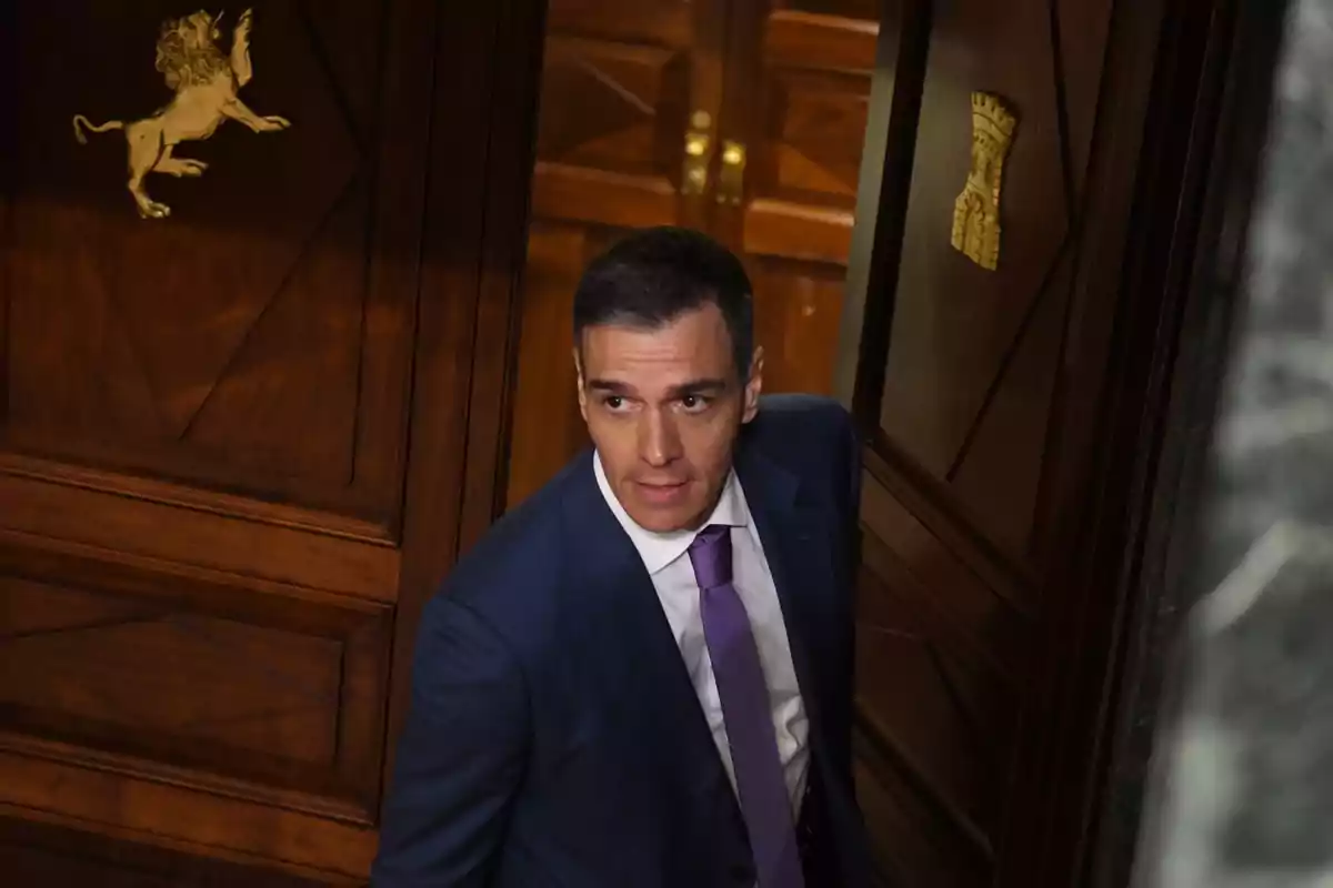 Hombre con traje azul y corbata morada entrando por una puerta de madera decorada con símbolos dorados