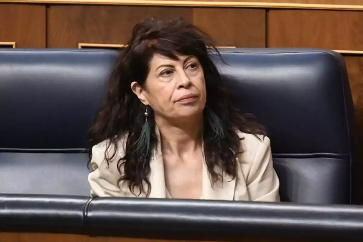Una mujer con cabello oscuro y rizado, sentada en un asiento de cuero azul en un entorno formal. Una mujer con cabello oscuro y rizado, sentada en un asiento de cuero azul en un entorno formal.