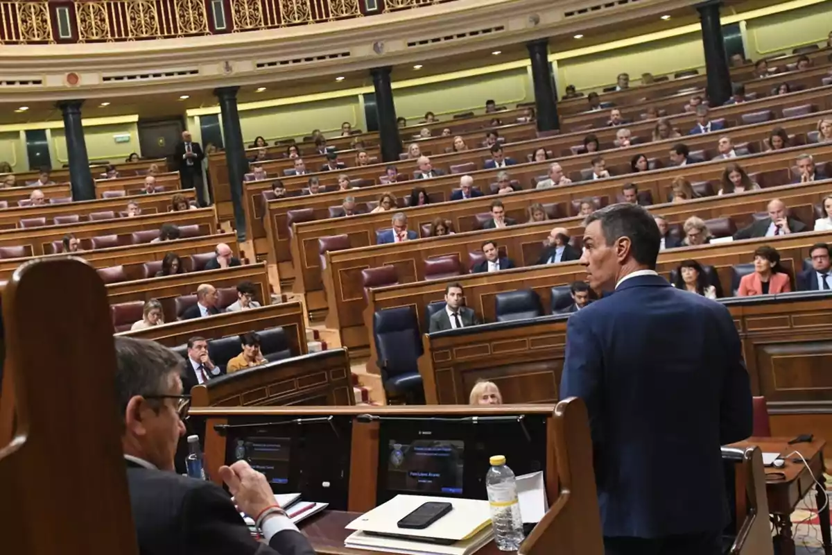 Una persona de pie en un podio frente a una audiencia en un gran salón con asientos de madera y detalles decorativos en el techo.