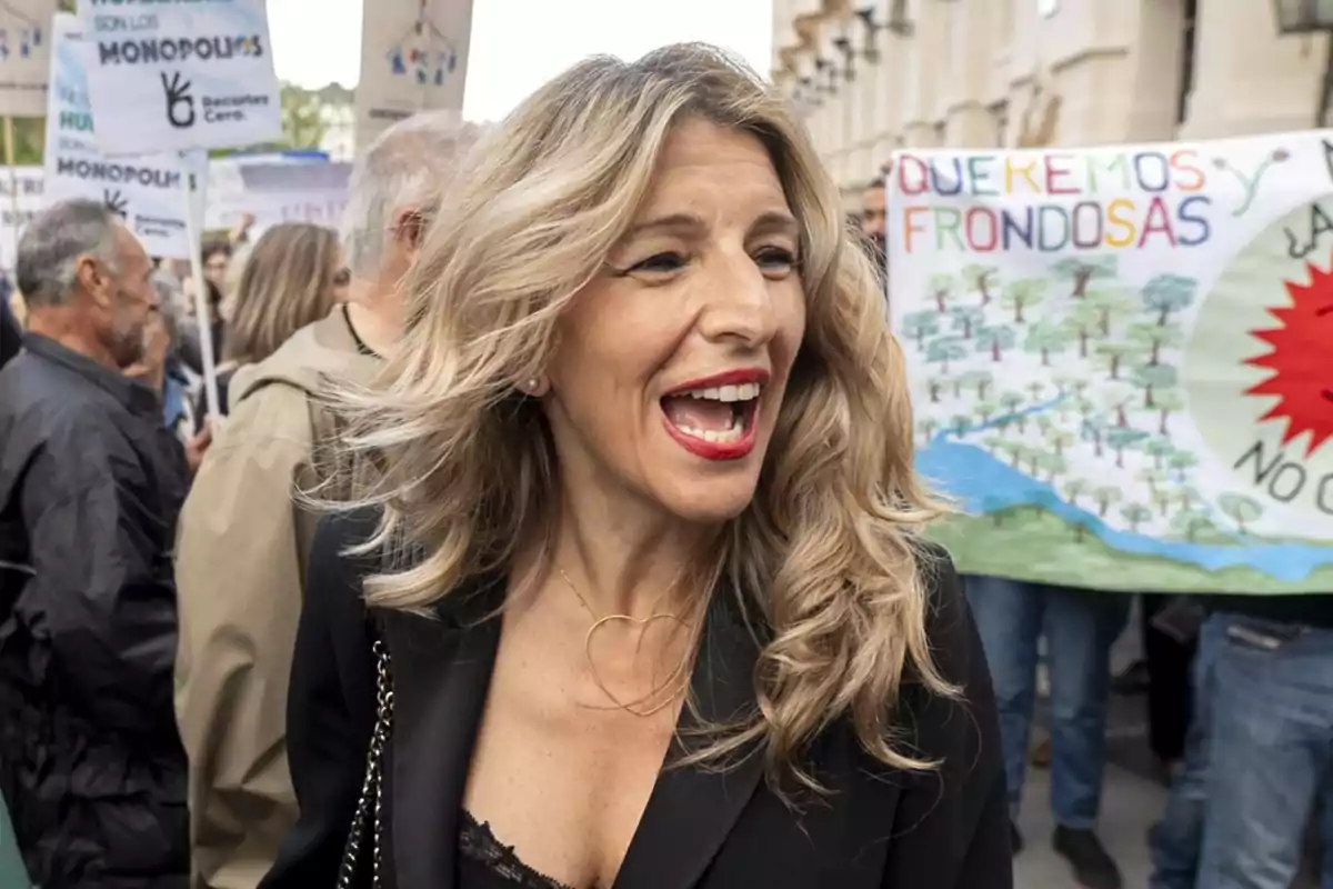 Una mujer sonriente con cabello rubio y labios rojos está en una manifestación rodeada de personas que sostienen pancartas. Una mujer sonriente con cabello rubio y labios rojos está en una manifestación rodeada de personas que sostienen pancartas.