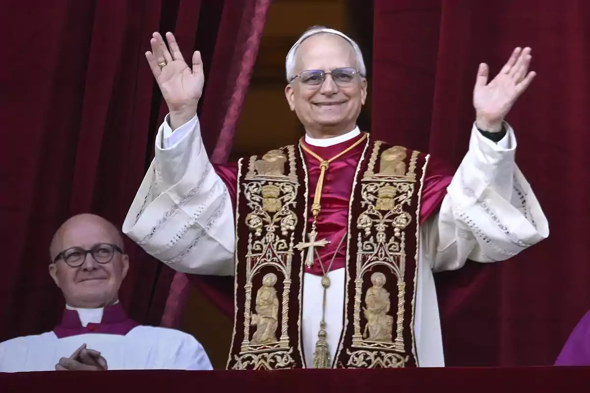 Un hombre con vestimenta religiosa levanta las manos en señal de saludo mientras otra persona lo observa sonriendo.