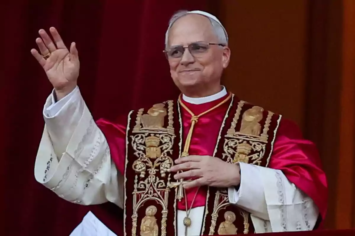 Un hombre vestido con vestimenta religiosa elaborada, incluyendo una sotana roja y una estola decorada, levanta la mano en un gesto de saludo frente a un fondo rojo. Un hombre vestido con vestimenta religiosa elaborada, incluyendo una sotana roja y una estola decorada, levanta la mano en un gesto de saludo frente a un fondo rojo.