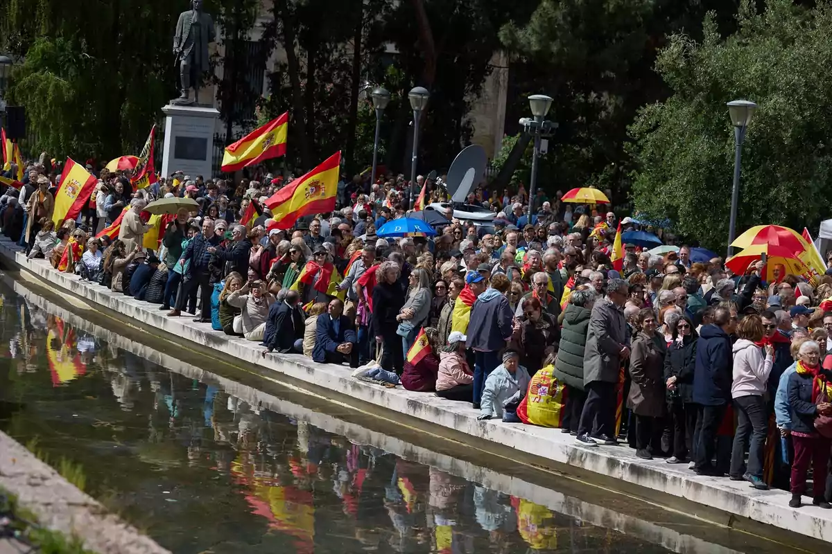Una multitud de personas se reúne junto a un estanque, muchas de ellas portando banderas de España y paraguas con los mismos colores, mientras una estatua se alza al fondo. Una multitud de personas se reúne junto a un estanque, muchas de ellas portando banderas de España y paraguas con los mismos colores, mientras una estatua se alza al fondo.