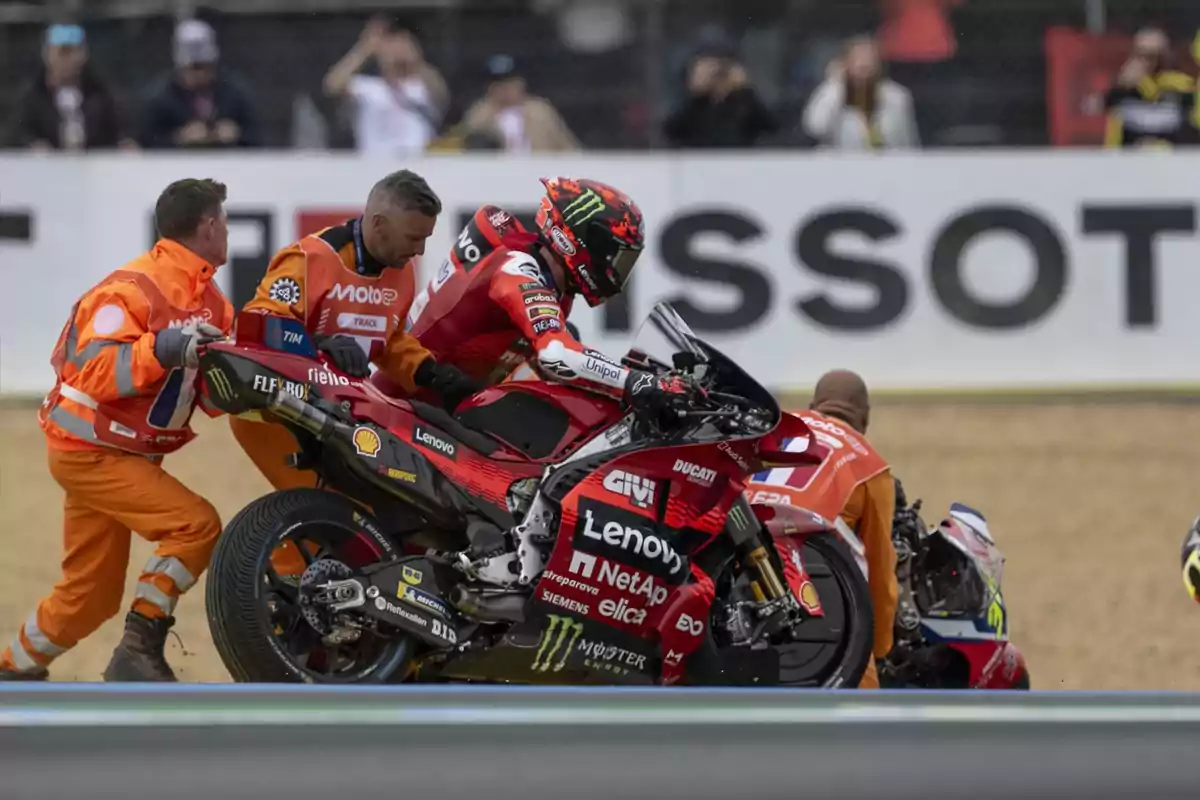 Un motociclista con traje rojo y casco está siendo asistido por personal de pista vestido de naranja mientras empujan una motocicleta Ducati en un circuito de carreras. Un motociclista con traje rojo y casco está siendo asistido por personal de pista vestido de naranja mientras empujan una motocicleta Ducati en un circuito de carreras.