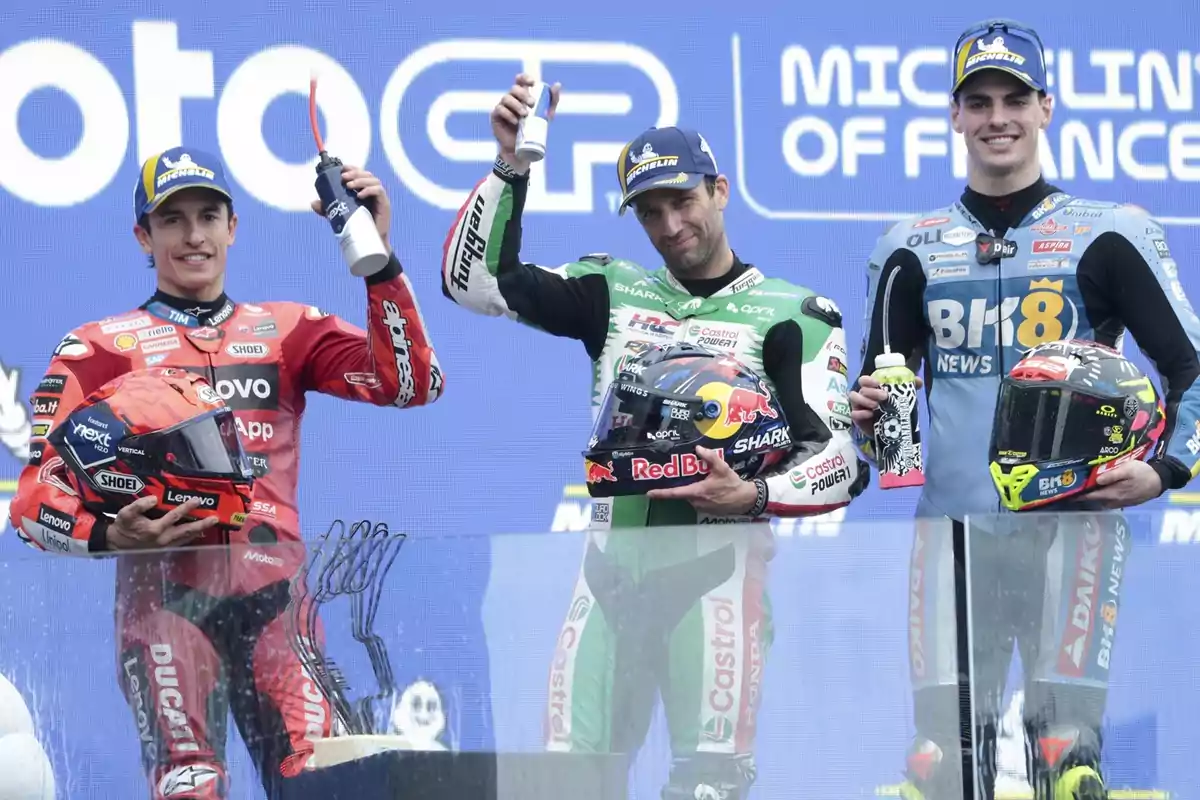 Tres pilotos de motociclismo en el podio celebran con trofeos y botellas, cada uno sosteniendo su casco, con un fondo que muestra el logotipo de MotoGP y Michelin.