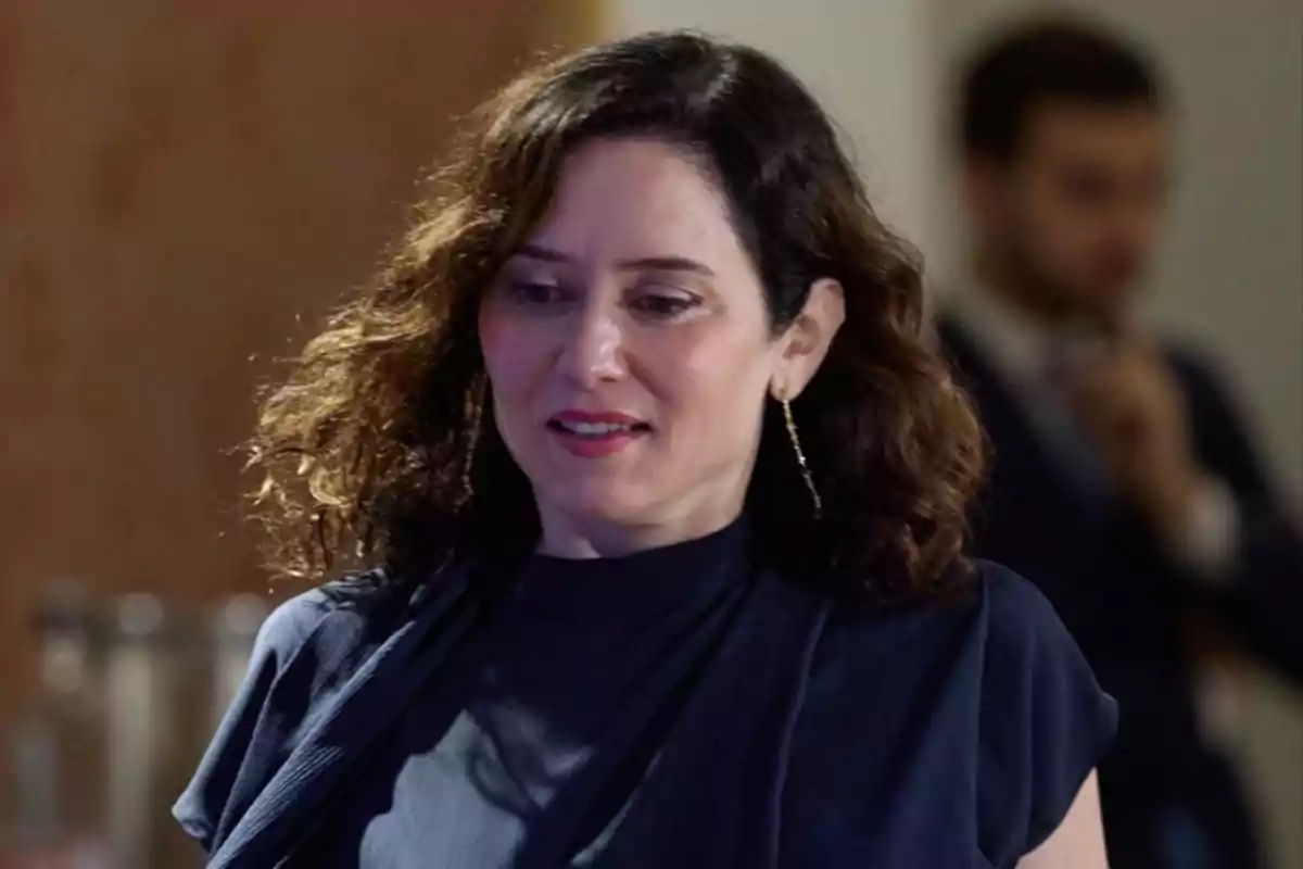 Una mujer con cabello rizado y pendientes largos, vestida de azul oscuro, en un entorno interior con una persona desenfocada al fondo. Una mujer con cabello rizado y pendientes largos, vestida de azul oscuro, en un entorno interior con una persona desenfocada al fondo.