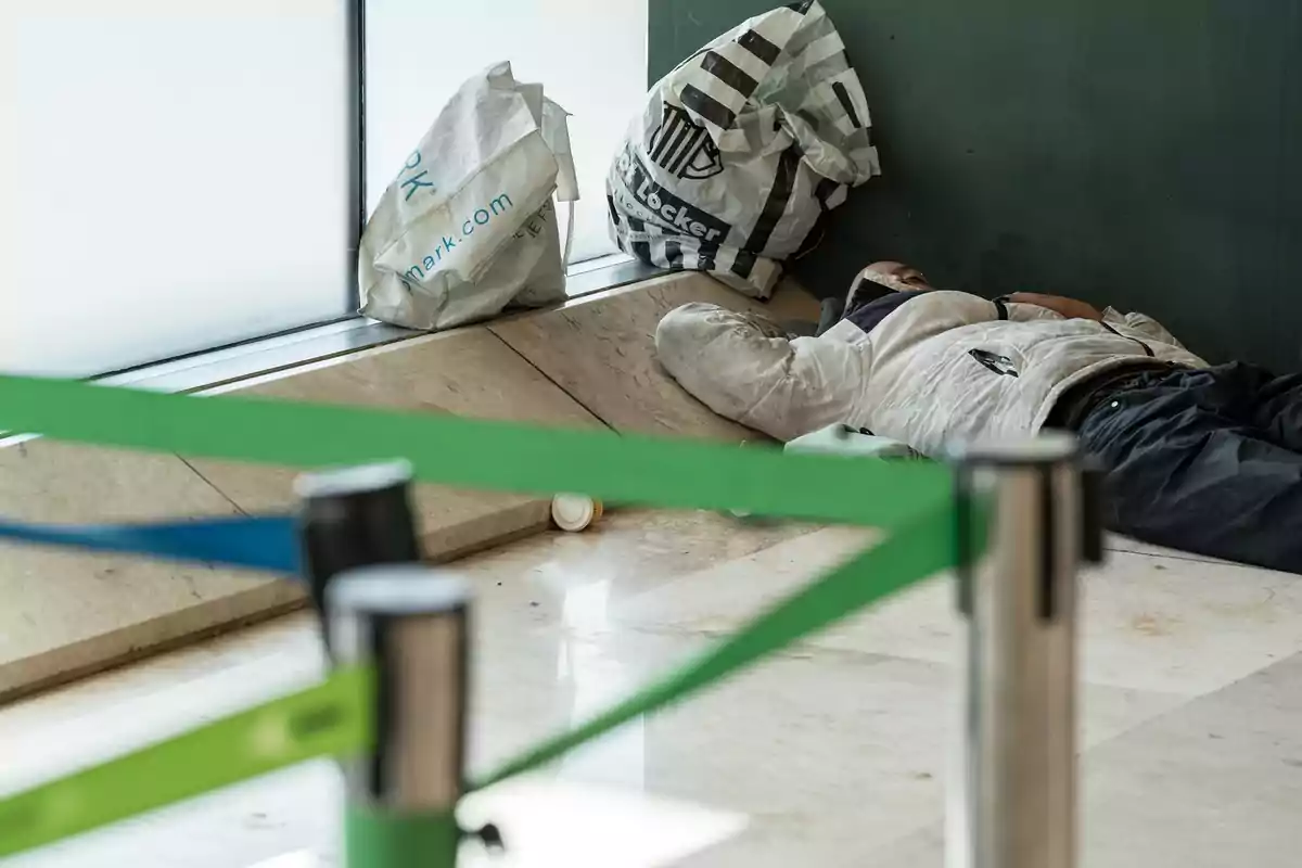 Una persona duerme en el suelo junto a una pared, con bolsas a su lado y cintas de seguridad en primer plano. Una persona duerme en el suelo junto a una pared, con bolsas a su lado y cintas de seguridad en primer plano.