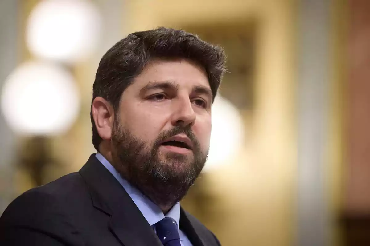 Hombre de cabello y barba oscuros vistiendo traje y corbata azul en un entorno interior con luces desenfocadas al fondo