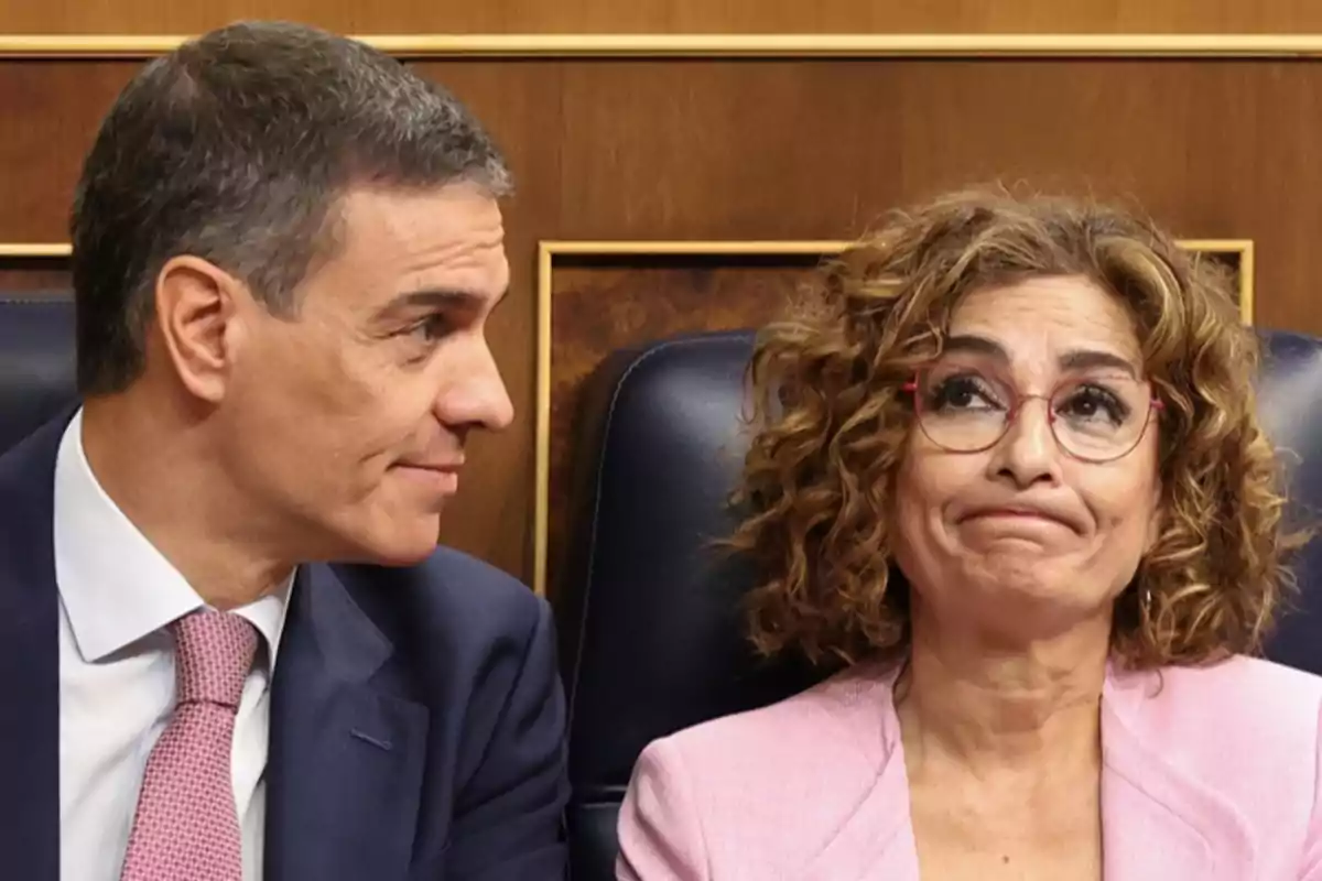 Dos personas sentadas en lo que parece ser un entorno formal, una de ellas mira a la otra que tiene una expresión facial de resignación o descontento.