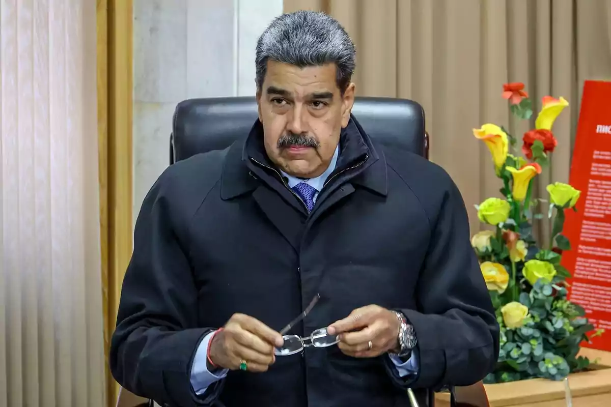 Un hombre con bigote y cabello canoso está sentado en una silla sosteniendo unas gafas, lleva un abrigo oscuro y una corbata azul, detrás hay cortinas y un arreglo de flores coloridas. Un hombre con bigote y cabello canoso está sentado en una silla sosteniendo unas gafas, lleva un abrigo oscuro y una corbata azul, detrás hay cortinas y un arreglo de flores coloridas.