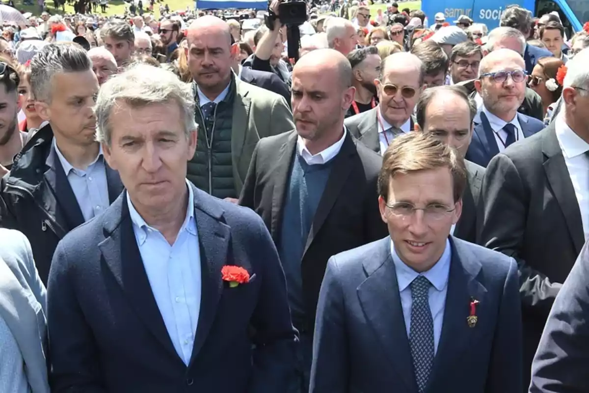 Un grupo de personas vestidas formalmente camina en una multitud al aire libre, algunos llevan claveles rojos en la solapa.