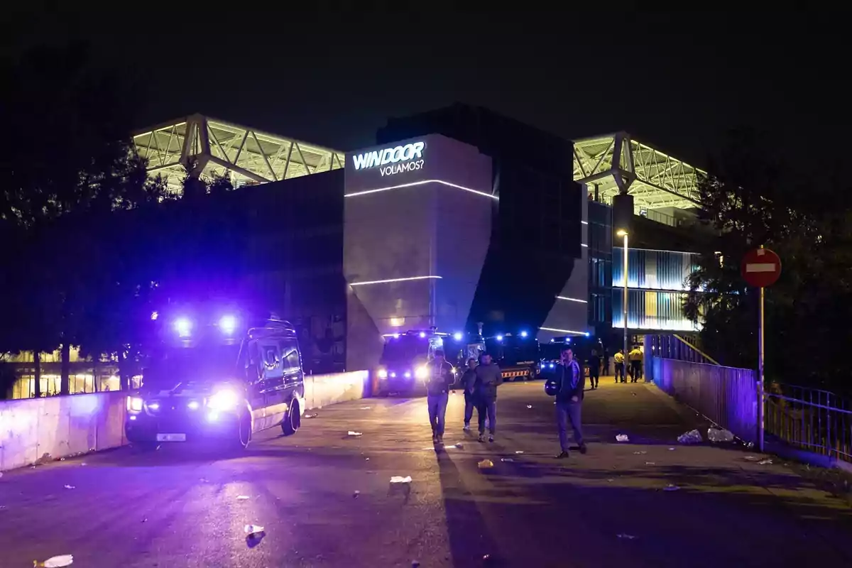Un edificio iluminado por la noche con varias personas y vehículos con luces azules en la calle.