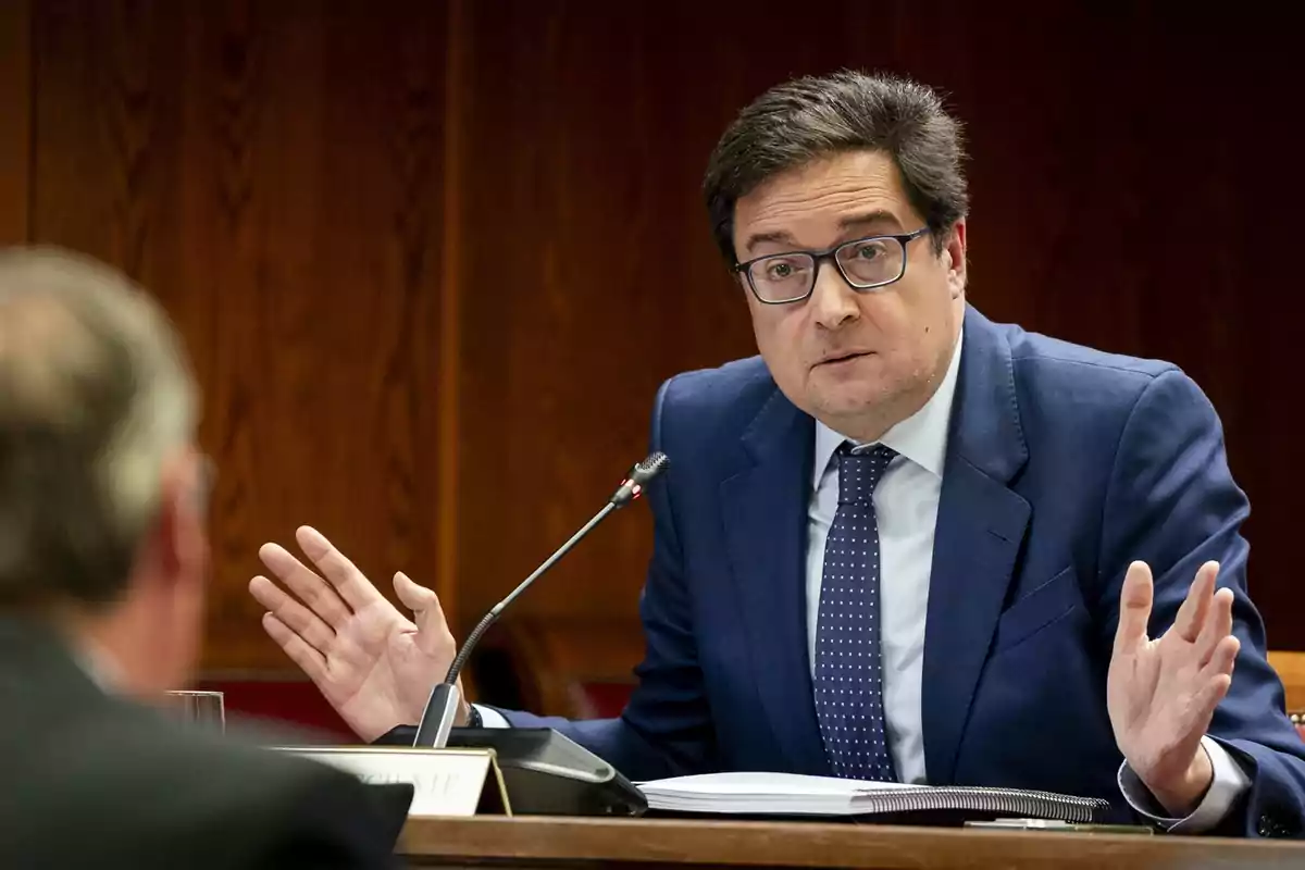 Hombre con gafas y traje azul hablando en una reunión formal con las manos levantadas y un micrófono frente a él Hombre con gafas y traje azul hablando en una reunión formal con las manos levantadas y un micrófono frente a él