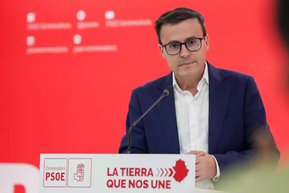 Hombre con gafas y traje azul hablando en un atril con fondo rojo y logotipos del PSOE de Extremadura