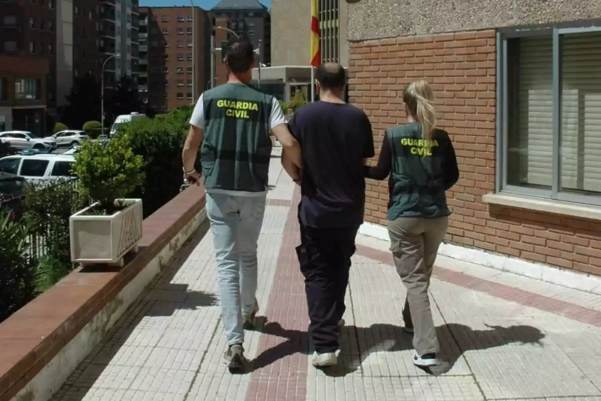 Dos agentes de la Guardia Civil escoltan a una persona por una acera en un entorno urbano.