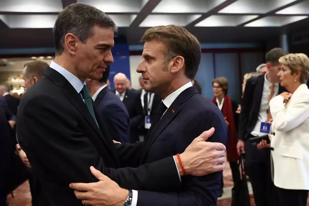 Dos hombres en traje se saludan con un abrazo en un evento formal rodeados de otras personas. Dos hombres en traje se saludan con un abrazo en un evento formal rodeados de otras personas.