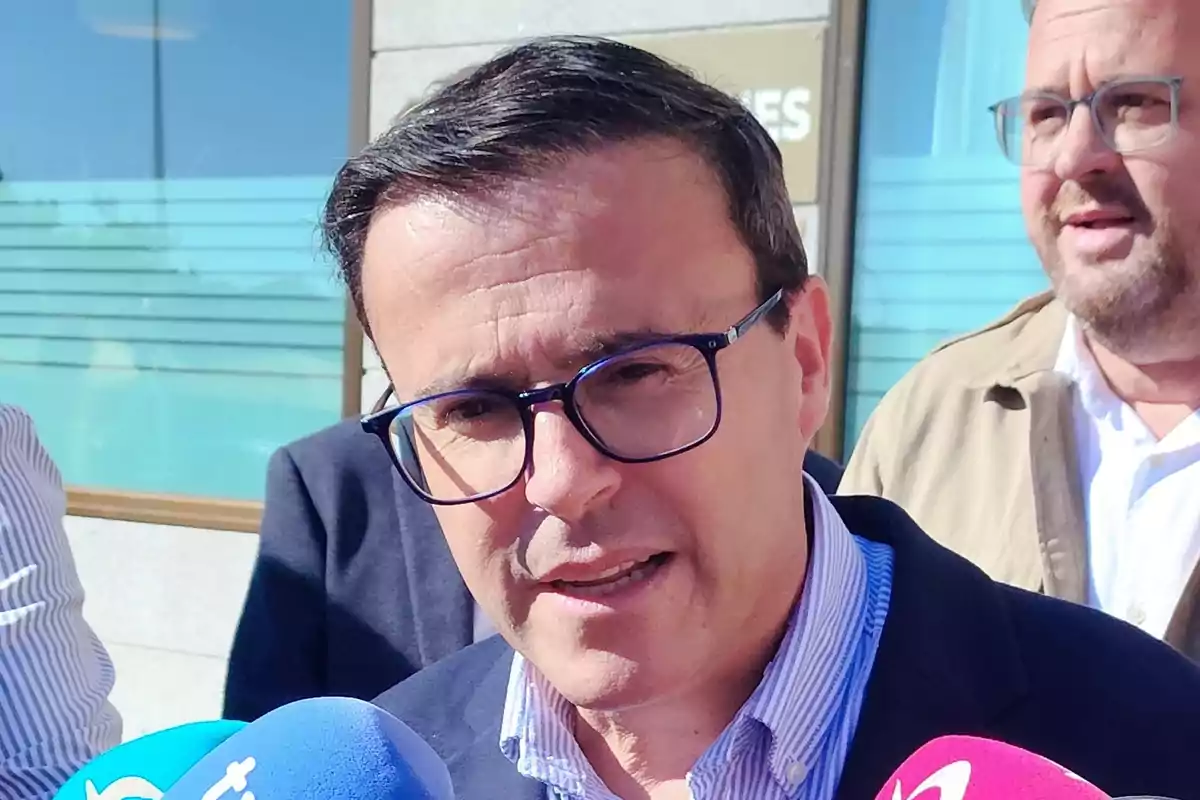Hombre con gafas hablando frente a varios micrófonos en un evento al aire libre Hombre con gafas hablando frente a varios micrófonos en un evento al aire libre