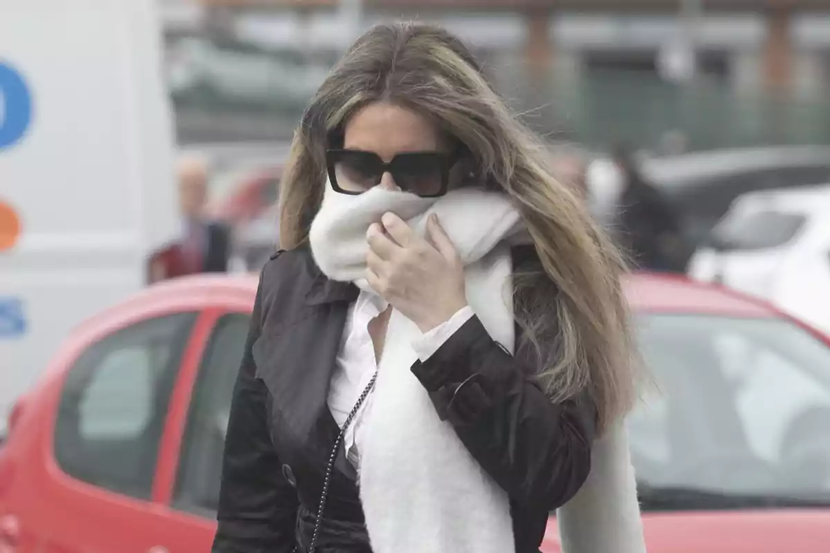 Mujer con gafas oscuras y bufanda blanca cubriéndose parte del rostro mientras camina frente a autos estacionados Mujer con gafas oscuras y bufanda blanca cubriéndose parte del rostro mientras camina frente a autos estacionados