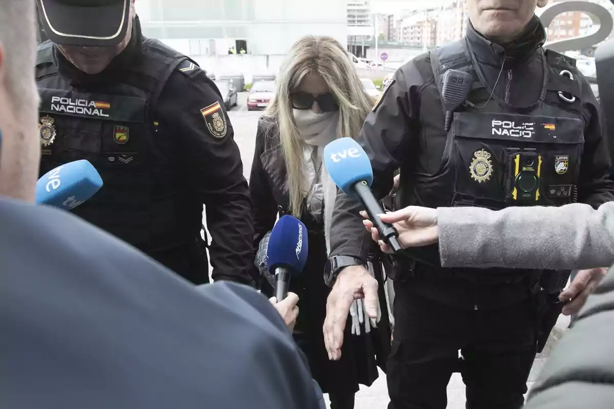 Una persona escoltada por dos agentes de policía mientras varios micrófonos de prensa se acercan para obtener declaraciones. Una persona escoltada por dos agentes de policía mientras varios micrófonos de prensa se acercan para obtener declaraciones.