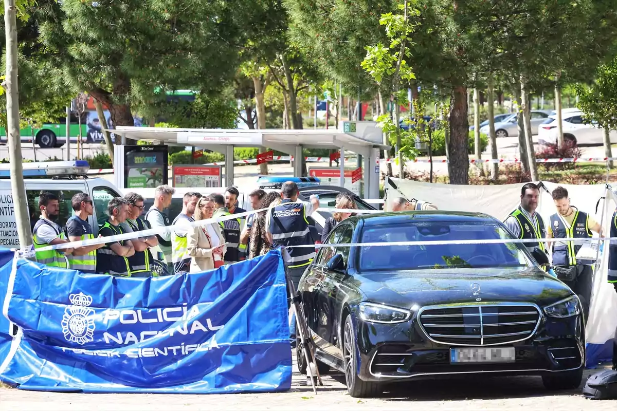 Un grupo de personas con chalecos de la policía se encuentra en una escena acordonada junto a un coche negro y una furgoneta, con un cartel de Un grupo de personas con chalecos de la policía se encuentra en una escena acordonada junto a un coche negro y una furgoneta, con un cartel de
