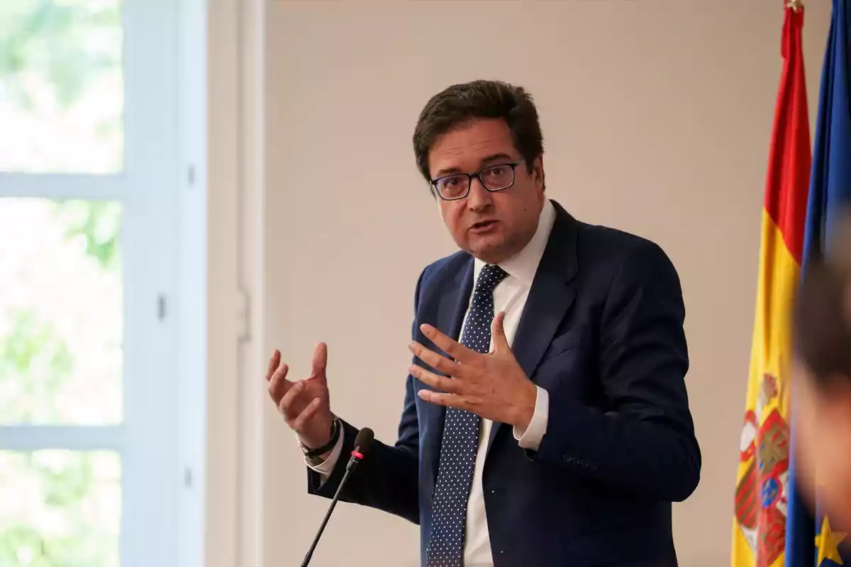 Hombre con gafas y traje azul hablando en una conferencia junto a una bandera de España y una bandera azul