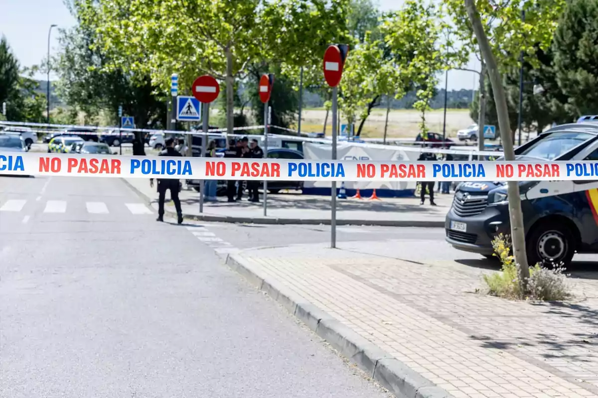 Una calle acordonada con cinta de la policía que dice "No pasar", con vehículos policiales y agentes al fondo.
