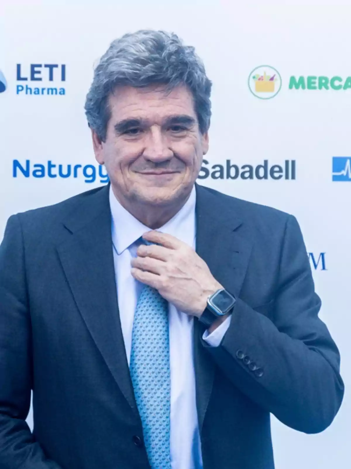 Hombre de cabello canoso con traje y corbata azul claro ajustándose la corbata frente a un fondo con logotipos de empresas Hombre de cabello canoso con traje y corbata azul claro ajustándose la corbata frente a un fondo con logotipos de empresas