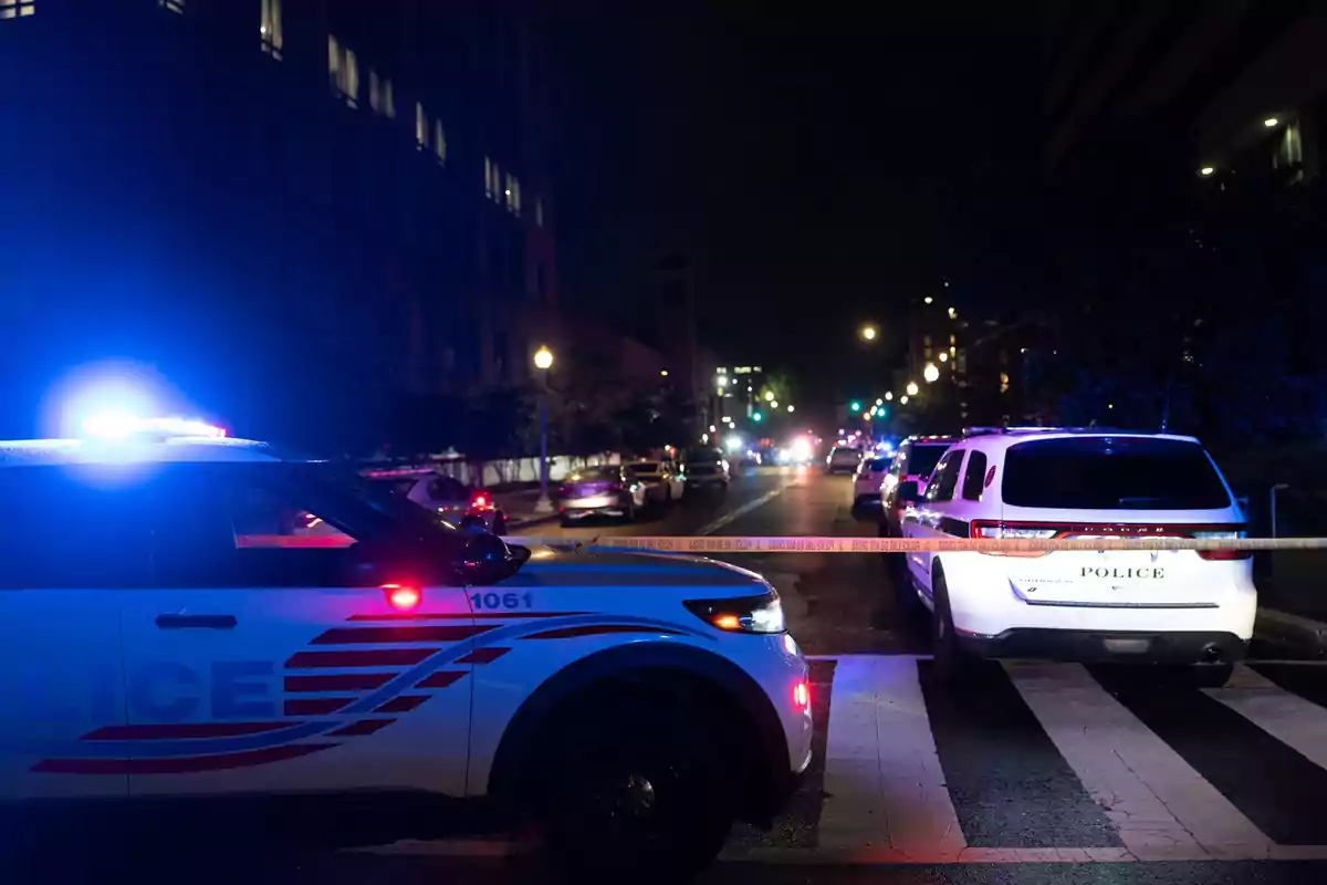 Una escena nocturna muestra vehículos policiales con luces encendidas bloqueando una calle con cinta de seguridad. Una escena nocturna muestra vehículos policiales con luces encendidas bloqueando una calle con cinta de seguridad.