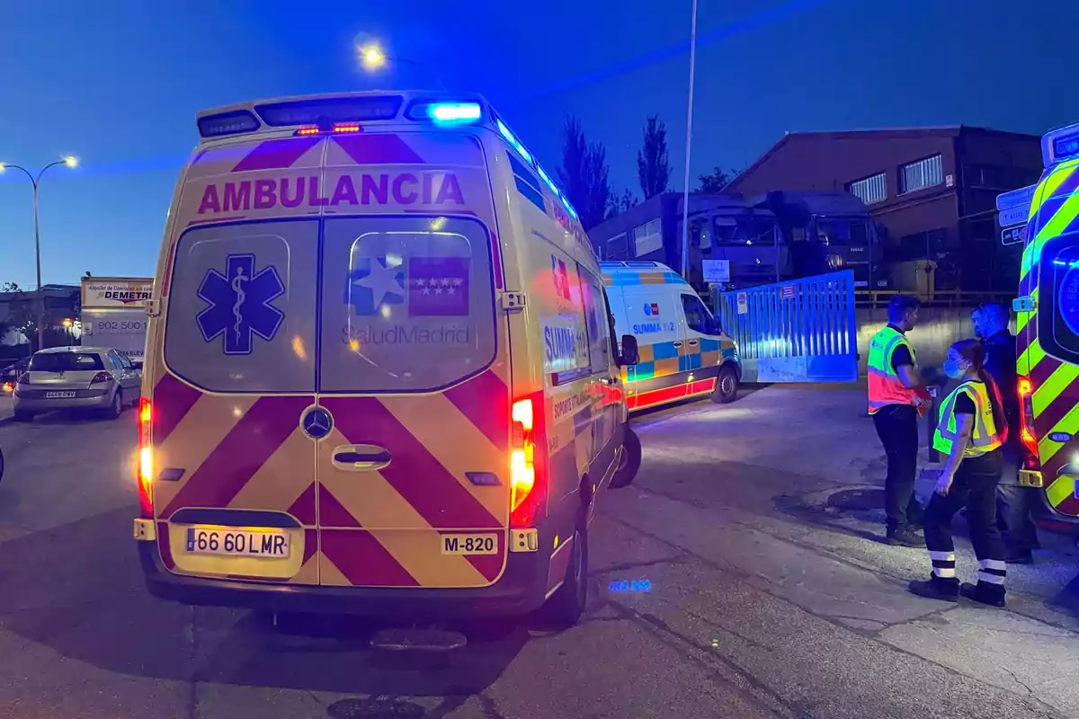 Una escena nocturna muestra varias ambulancias estacionadas en una calle, con luces de emergencia encendidas y personal de emergencia conversando cerca de ellas.