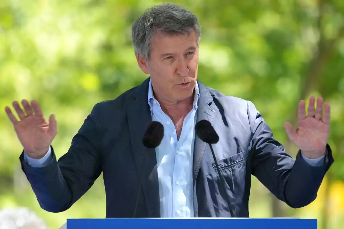 Hombre de cabello canoso hablando al aire libre frente a dos micrófonos con las manos levantadas y fondo de árboles desenfocado