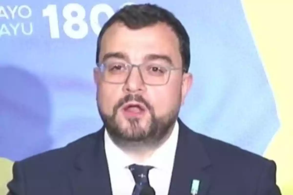 Hombre con barba y gafas hablando frente a un micrófono con fondo azul y amarillo Hombre con barba y gafas hablando frente a un micrófono con fondo azul y amarillo