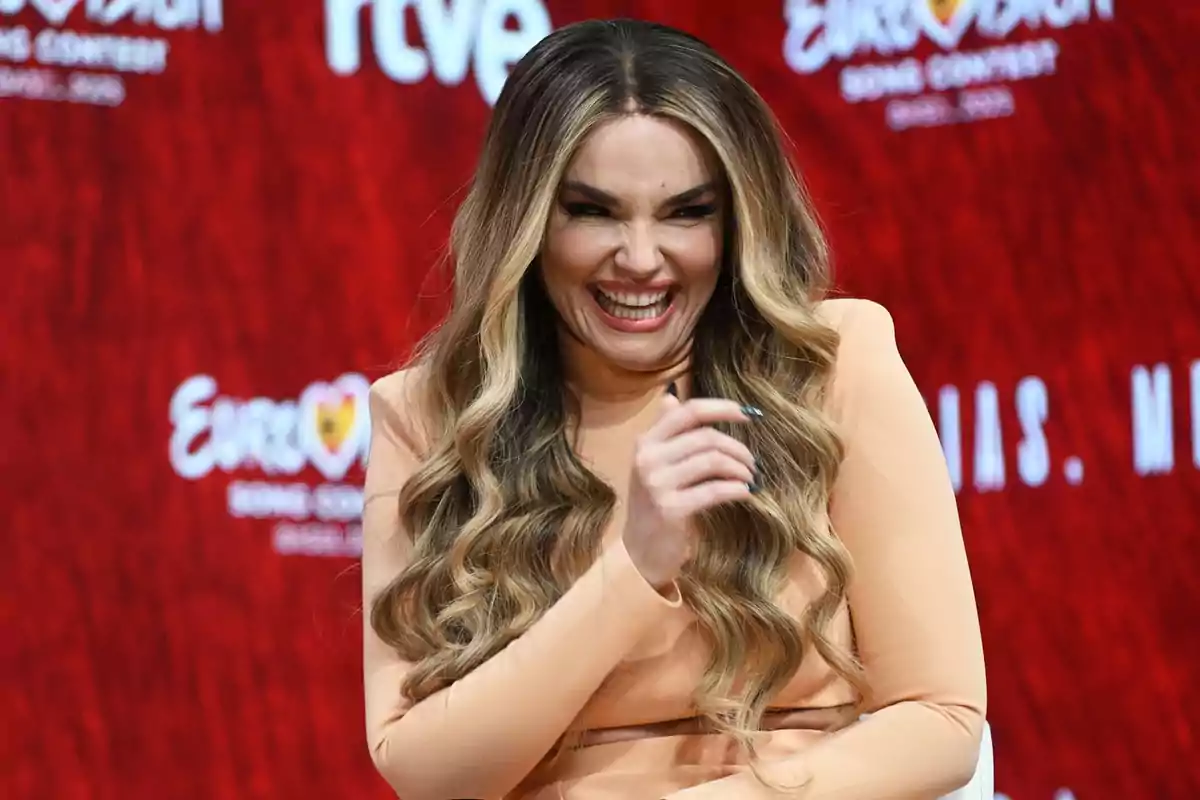 Mujer con cabello largo y ondulado sonríe ampliamente mientras está sentada frente a un fondo rojo con logotipos de Eurovision Mujer con cabello largo y ondulado sonríe ampliamente mientras está sentada frente a un fondo rojo con logotipos de Eurovision
