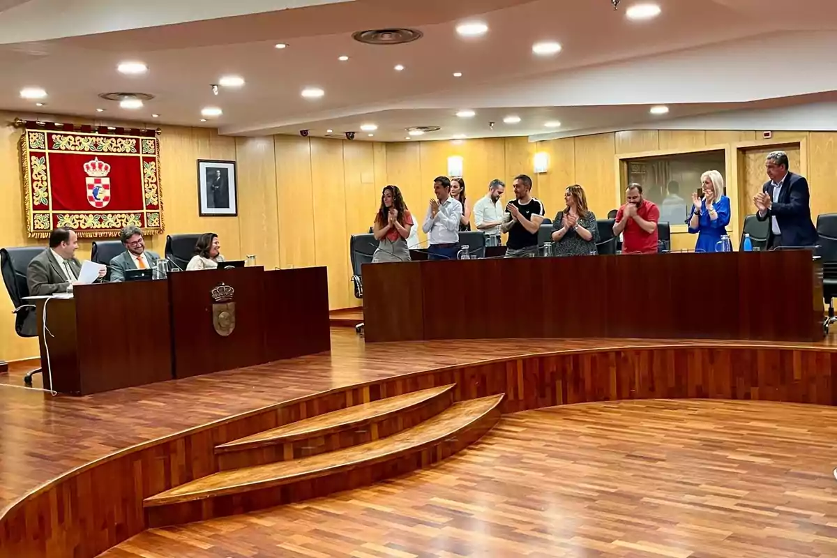 Personas en una sala de reuniones con paneles de madera, algunas sentadas y otras de pie aplaudiendo, con un escudo y una fotografía en la pared.
