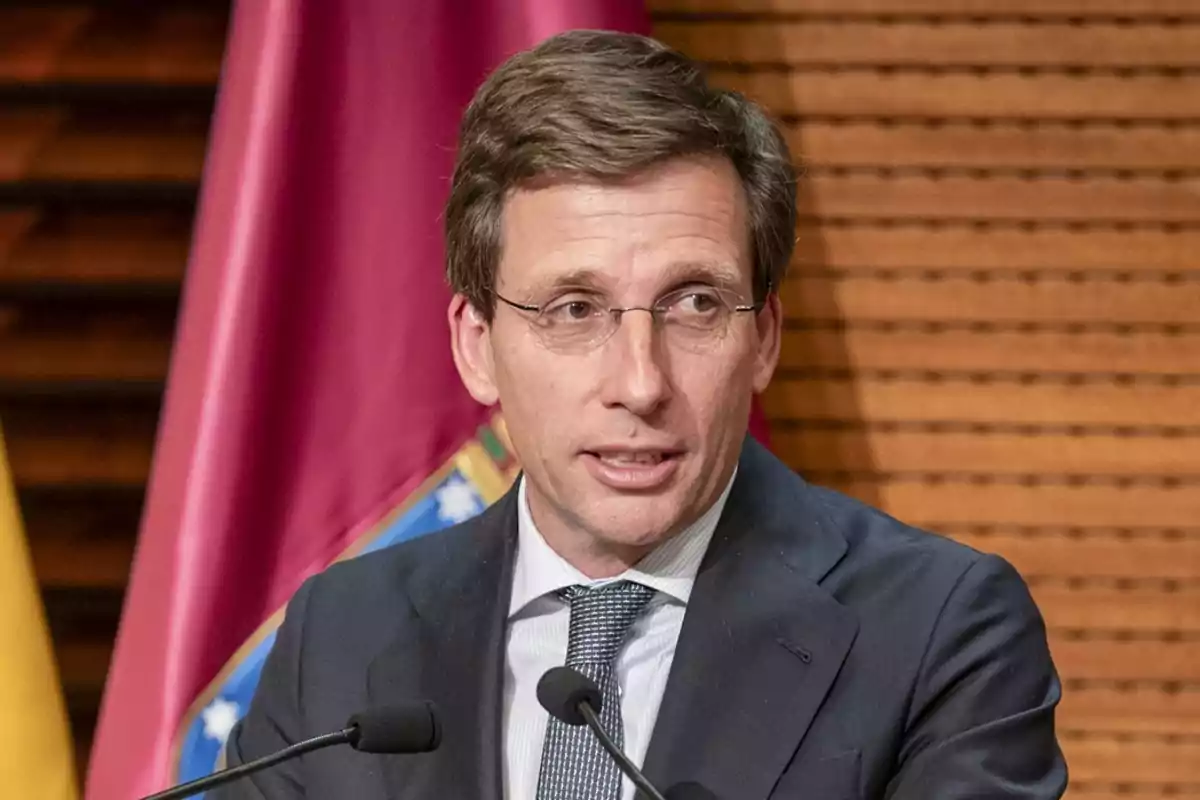 Hombre con gafas y traje hablando frente a micrófonos con una bandera de fondo Hombre con gafas y traje hablando frente a micrófonos con una bandera de fondo