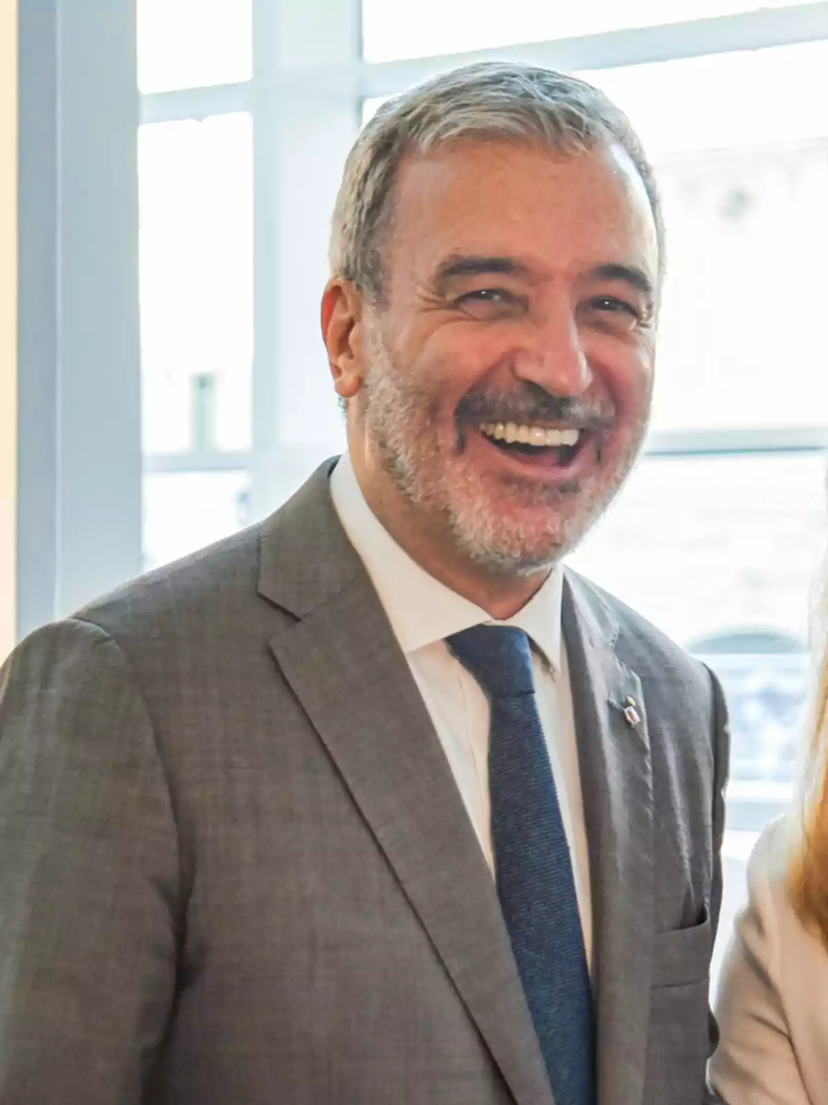 Hombre de cabello canoso y barba, vestido con traje gris y corbata azul, sonriendo frente a una ventana Hombre de cabello canoso y barba, vestido con traje gris y corbata azul, sonriendo frente a una ventana