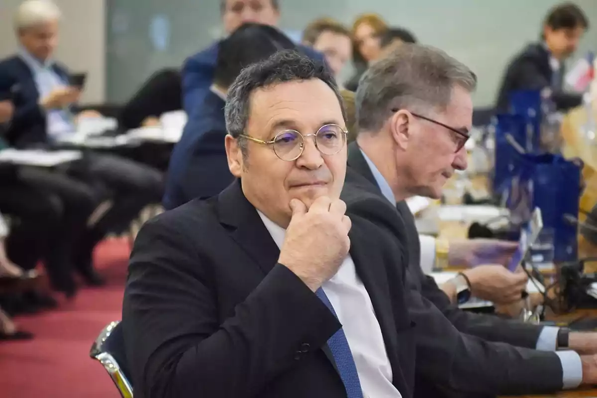 Hombre con gafas y traje oscuro sentado en una sala de conferencias rodeado de otras personas