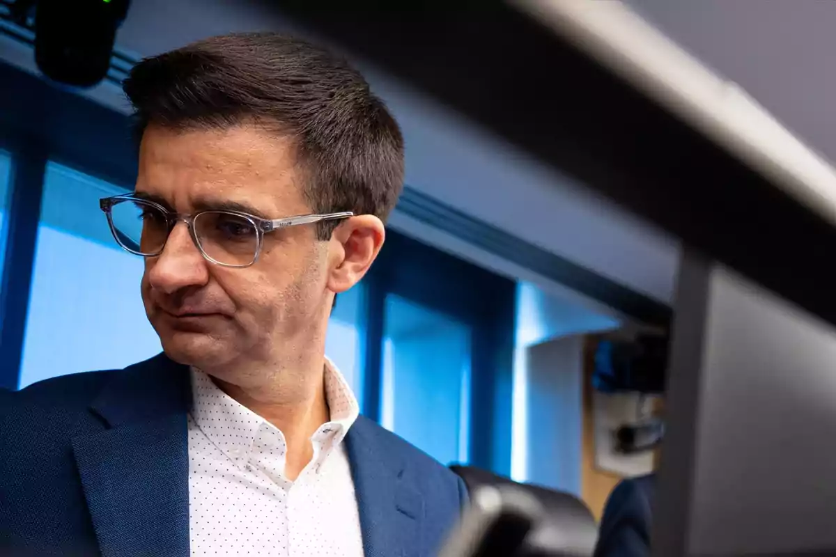 Hombre con gafas y saco azul mirando hacia abajo en una oficina moderna con ventanas grandes al fondo Hombre con gafas y saco azul mirando hacia abajo en una oficina moderna con ventanas grandes al fondo