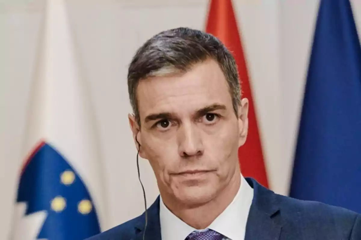 Hombre de cabello corto y canoso con auricular, vistiendo traje y corbata, frente a banderas de fondo. Hombre de cabello corto y canoso con auricular, vistiendo traje y corbata, frente a banderas de fondo.