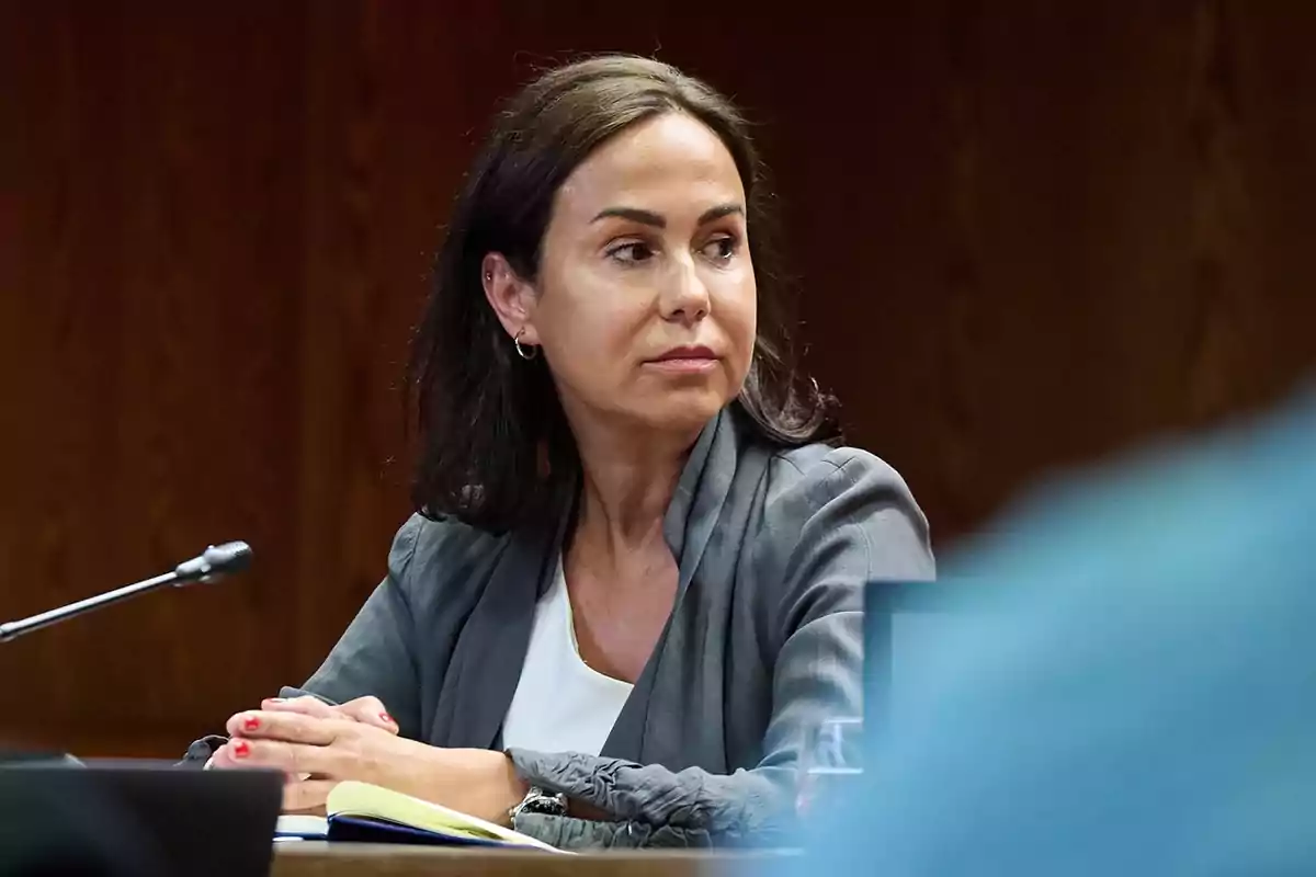 Mujer de cabello oscuro sentada en una mesa con un micrófono y una libreta, mirando hacia un lado en un entorno formal Mujer de cabello oscuro sentada en una mesa con un micrófono y una libreta, mirando hacia un lado en un entorno formal