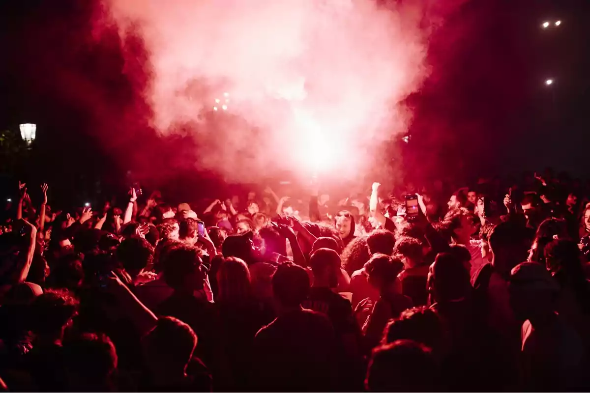 Multitud de personas reunidas de noche celebrando con una bengala roja que ilumina el ambiente y genera humo