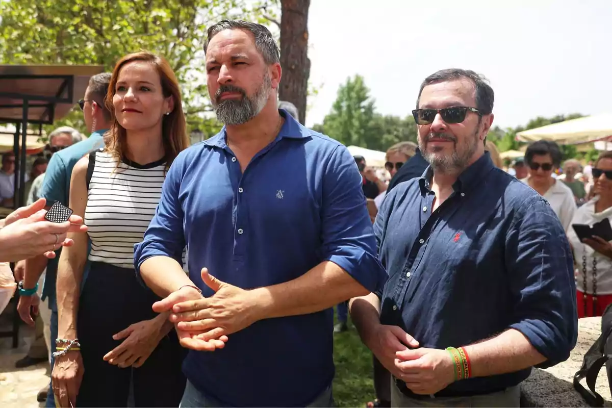 Tres personas de pie en un evento al aire libre rodeadas de más gente en un día soleado