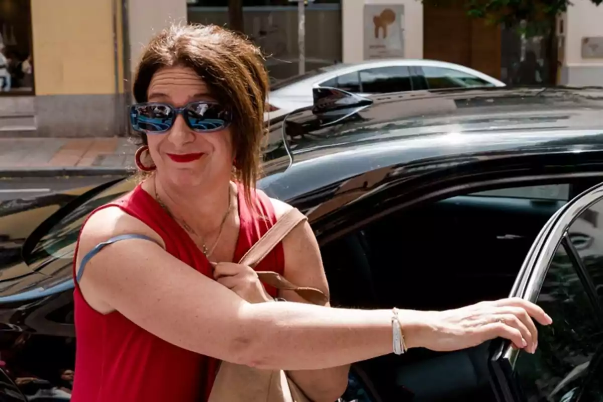 Mujer con vestido rojo y gafas de sol sonriendo mientras abre la puerta de un coche negro en una calle urbana