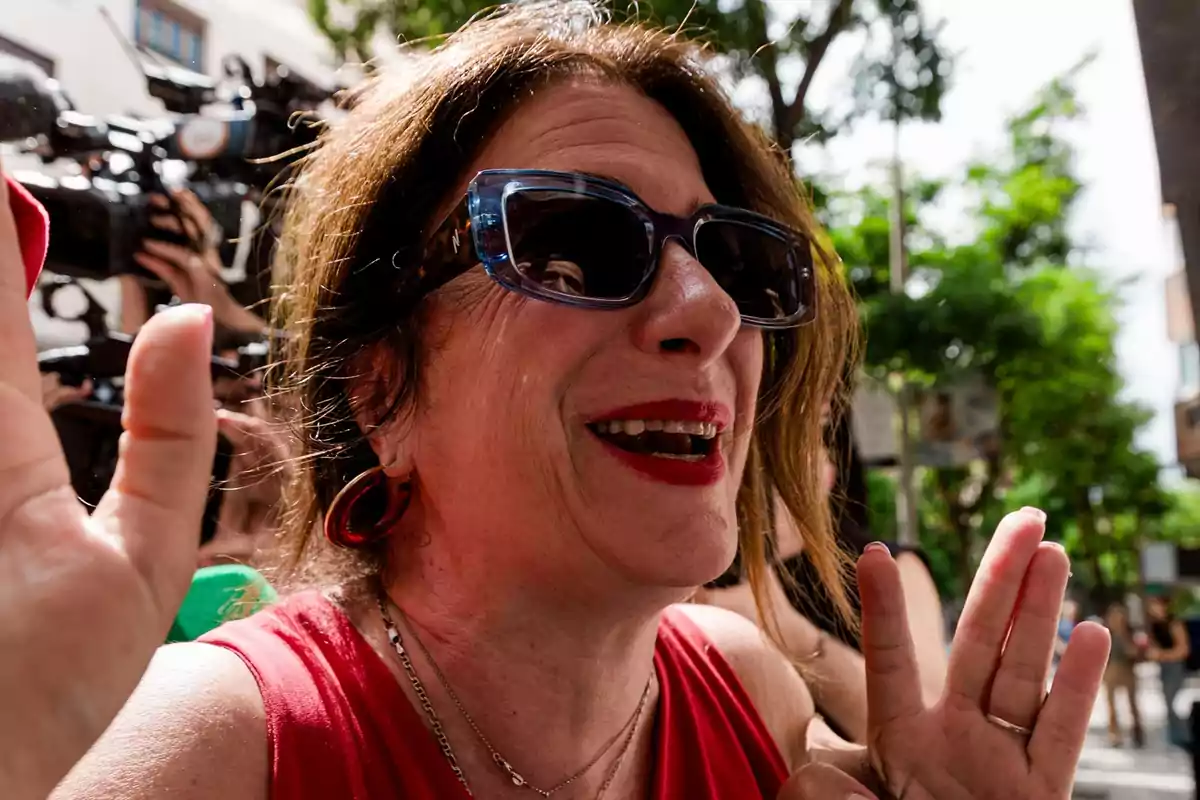 Mujer sonriente con gafas de sol y labios rojos rodeada de cámaras y personas al aire libre Mujer sonriente con gafas de sol y labios rojos rodeada de cámaras y personas al aire libre
