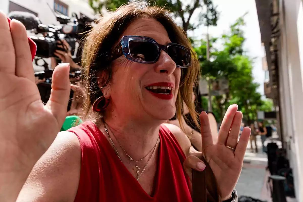 Mujer sonriente con gafas de sol y vestido rojo levantando las manos mientras es fotografiada en la calle Mujer sonriente con gafas de sol y vestido rojo levantando las manos mientras es fotografiada en la calle
