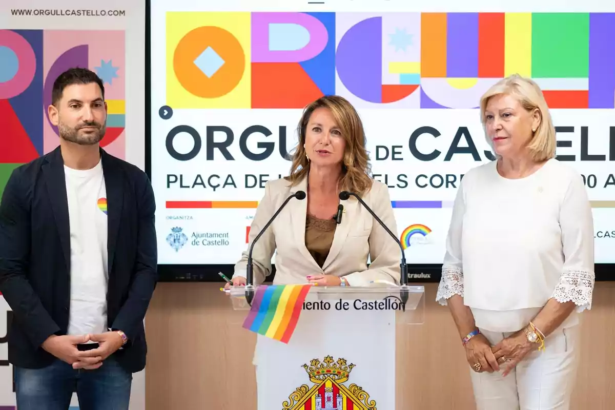 Tres personas participan en una rueda de prensa sobre el Orgullo en Castellón, con una bandera arcoíris en el atril y un cartel colorido de fondo