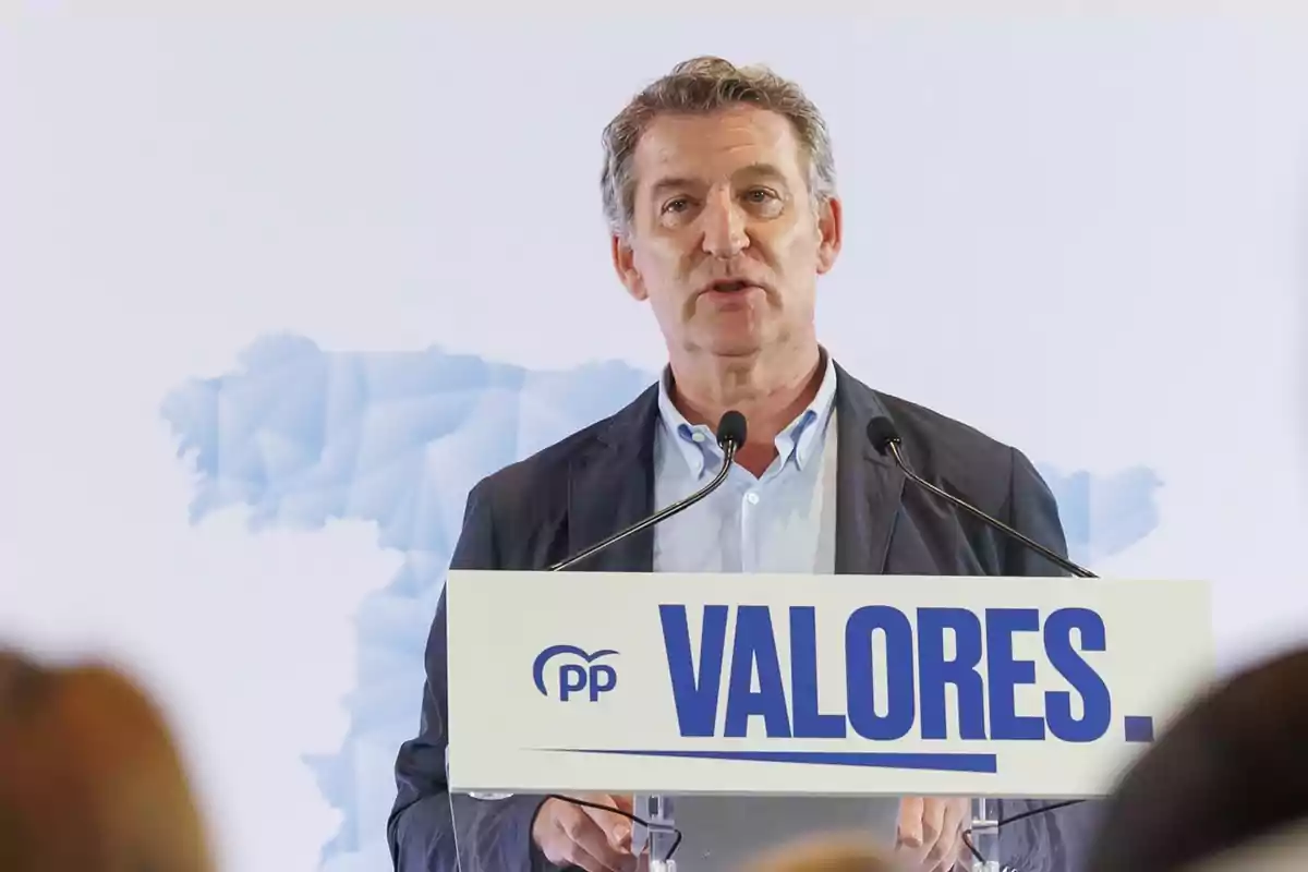 Hombre hablando en un atril con el logo del PP y la palabra VALORES en grande, fondo con mapa de España difuminado. Hombre hablando en un atril con el logo del PP y la palabra VALORES en grande, fondo con mapa de España difuminado.