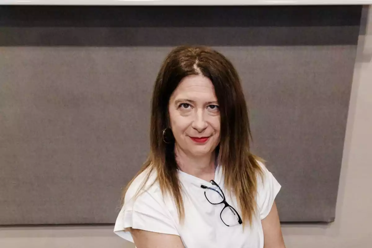 Mujer de cabello castaño lacio y largo con camiseta blanca y gafas colgadas en el cuello, sentada frente a un fondo gris, mirando a la cámara con expresión seria. Mujer de cabello castaño lacio y largo con camiseta blanca y gafas colgadas en el cuello, sentada frente a un fondo gris, mirando a la cámara con expresión seria.